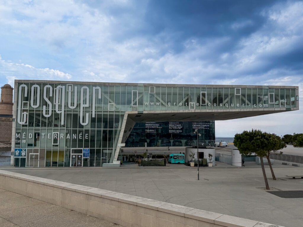 The image displays the Cosquer Méditerranée interpretation center, a museum located in Marseille, France. Function: It houses a detailed reconstruction of the Cosquer Cave, a submerged Paleolithic cave containing prehistoric rock art. Architecture: The building is situated in the harbor area next to the MuCEM and is known for its modern, architectural design. Experience: Visitors can take a tour of the replica cave using autonomous vehicles